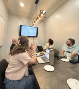 Blue Canopy Marketing team members gather around a meeting table during an April team meeting, reviewing information displayed on a television screen while discussing marketing, web development, and industry initiatives.